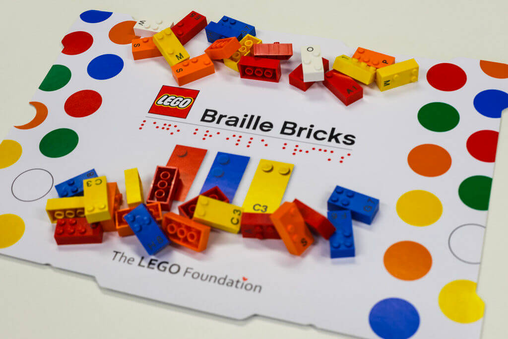 Coperchio della scatola LEGO Braille, con su scritto Braille Bricks in caratteri latini e Braille. Alcuni mattoncini colorati con sia scrittura Braille e latina.