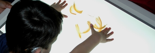 Bambino sordocieco gioca con delle piume di uccello gialle poste sopra uno schermo luminoso.